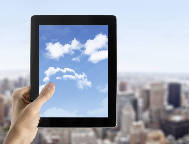 A photo of someone's hand holding a tablet device looking across a cityscape with bluesky and clouds in focus